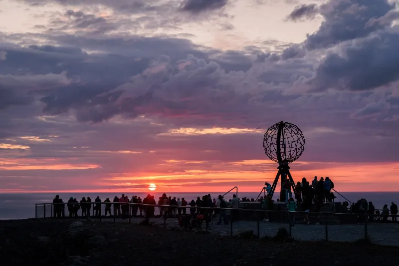 Il globo di Capo Nord di notte contro un cielo rosa e oro, persone che guardano il sole basso sul mare