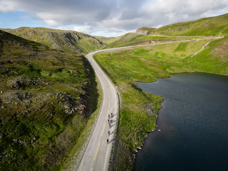 Una strada che serpeggia sulle colline verdi di Magerøya accanto a un lago scuro, qualche rider sulla strada