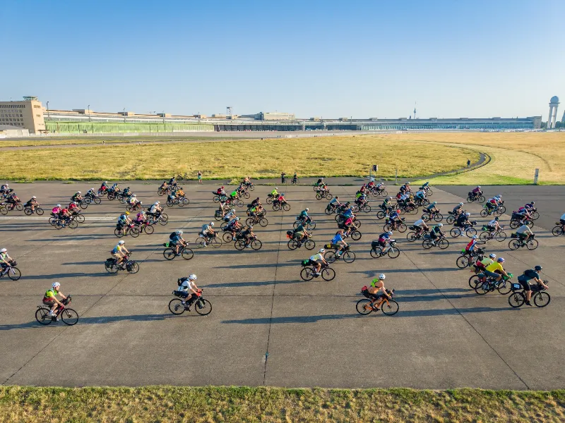 Decine di rider attraversano la pista di Tempelhof alla partenza di Berlino, sullo sfondo il vecchio terminal